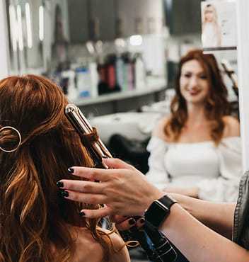 Person with red hair getting a hairstyle with a curling iron in a salon.