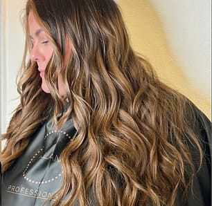 Woman with long, wavy, light brown hair in a salon cape, facing sideways against a beige background.