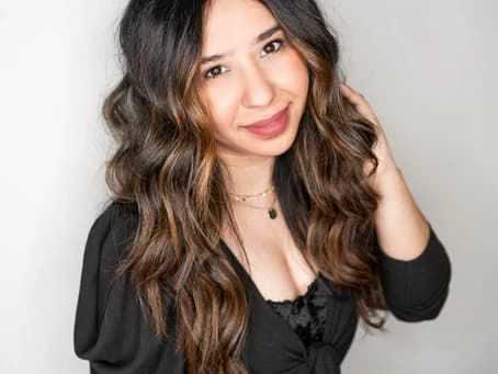 Young woman with long wavy hair, smiling and touching her hair, wearing a black top.