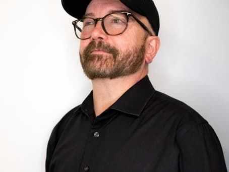 Man in black shirt and cap with glasses, looking up against a plain background.