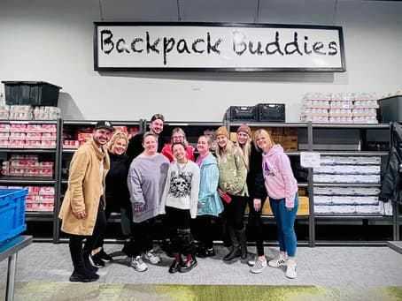 Group of people smiling in front of Backpack Buddies sign and shelves with supplies.