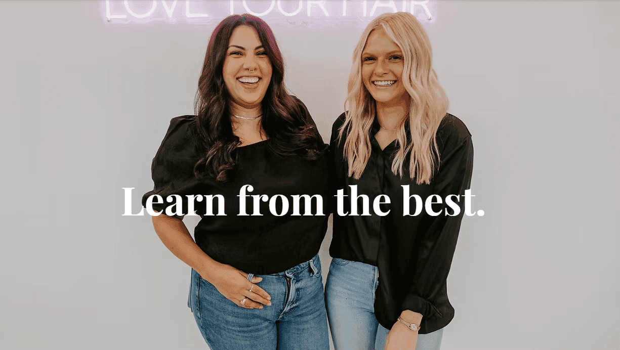 Two smiling women in black tops with a sign reading "Learn from the best" in front of them.
