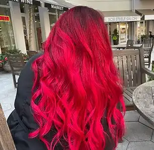 Person with long, vibrant red hair sitting on a wooden chair in an outdoor shopping area.
